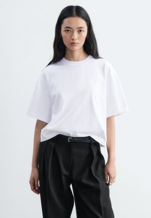 Un mannequin aux longs cheveux foncés se tient devant un fond clair, vêtu d’un T-shirt blanc oversize, d’un pantalon noir et d’une ceinture noire.