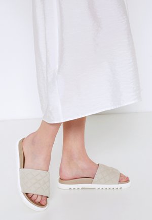 Person trägt beige gesteppte Schlupfsandalen mit weißen Sohlen und weißem knöchellangem Kleid und steht auf einem weißen Boden.