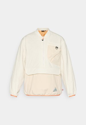 Jasno-beżowy pikowany pulower z regulowanym półzamkiem, kieszenią z przodu oraz pomarańczowymi akcentami na mankietach i dole.