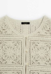 Krämfärgad virkad cardigan med cirkulära blommönster, rund halsringning och knäppning med tre knappar framtill.