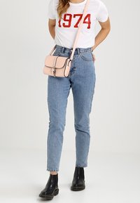 Lyserød crossbody taske med klap og sølv hardware, båret over en hvid t-shirt med rød tekst, parret med højtaljede blå jeans og sorte støvler.