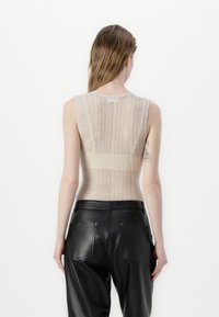 Lahka bež majica brez rokavov z teksturiranim pletenim dizajnom, kombinirana s črnimi usnjenimi hlačami, ki imajo zadnje žepe in gladko površino.