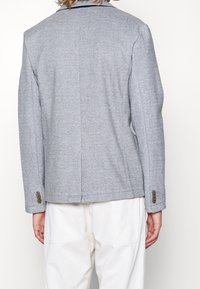 Persona vistiendo un blazer gris con puños abotonados y pantalones blancos, vista desde atrás sobre un fondo blanco liso.