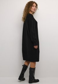 Juoda megzta suknelė su laisvu siluetu, ilgomis rankovėmis ir banguotais rankogaliais. Lygus audinys ir kelių ilgio dizainas. Sujungta su juodais aukštakulniais batais iki kelių.