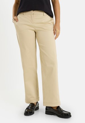 Pantalon beige à jambes larges porté avec des mocassins noirs et un haut noir, une main dans la poche et l'autre reposant sur le côté.