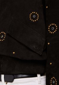 Veste en daim noire avec des clous dorés circulaires et des ornements de pierres bleues sur tissu noir côtelé, associée à un pantalon blanc et une ceinture noire.