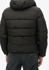 Musta toppatakki hupulla, jossa vaakasuora tikattu kuvio, kirjailtu logo takana ja sileä pinta.