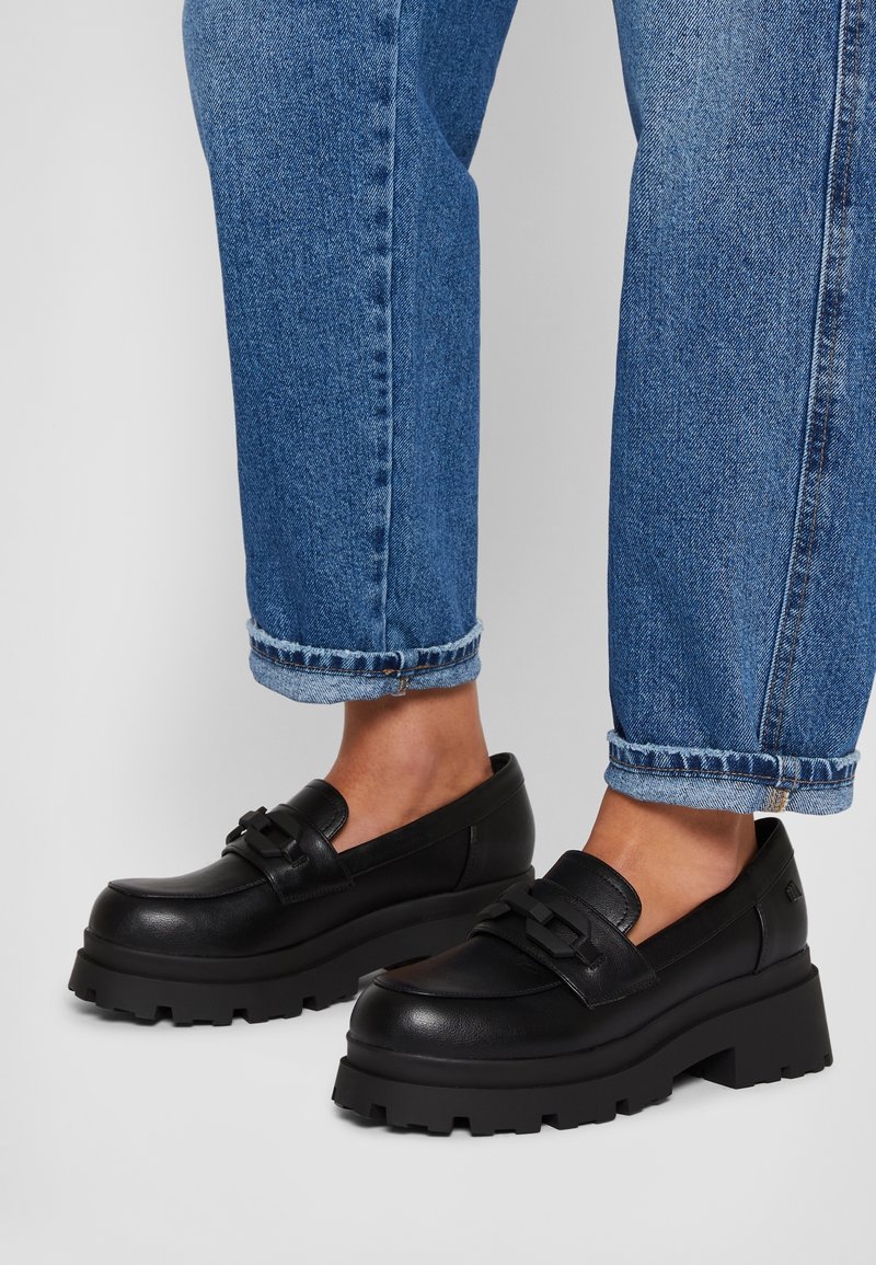 Mocassins noirs à plateforme épaisse portés avec un jean en denim bleu retroussé sur une surface gris clair.
