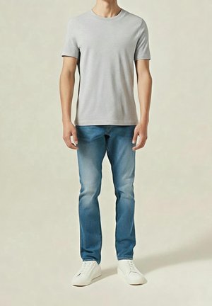 Mann trägt ein schlichtes hellgraues T-Shirt mit kurzen Ärmeln, blaue Jeans und weiße Sneaker, steht vor einem neutralen Hintergrund.
