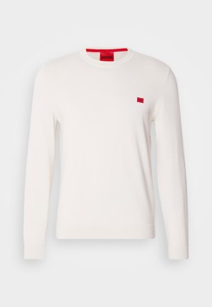 Bela majica s kružnim izrezom i dugimi rokavi, izdelana iz mehkega materiala. Ima majhno rdečo oznako na prsih in kontrastno rdečo etiketo na vratu.