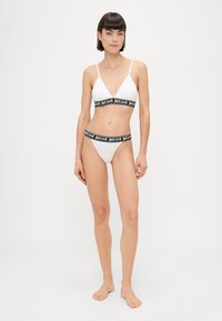 Balts bikini komplekts, kas sastāv no trīsstūra formas krūštura ar regulējamiem siksniņām un melna josla, apvienots ar atbilstošiem augstiem apakšveļas gabaliem.