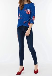 Blusa floreale blu con maniche a tre quarti, abbinata a jeans skinny neri e scarpe col tacco rosse. Tessuto morbido, motivi vivaci.