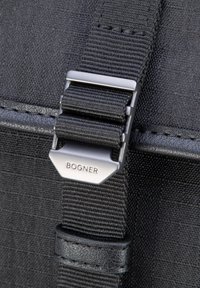 Detajl črne tkanine torbe z metalno zaponko z napisom "BOGNER" in teksturiranim najlonskim trakom. Gladek usnjen poudarek blizu zaponke.