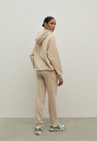 Béžová mikina s kapucňou, šnúrkou a klokankou, v kombinácii s ladnými teplákmi. Vyznačuje sa jemnou textúrou a rebrovanými manžetami. Strieborné tenisky.