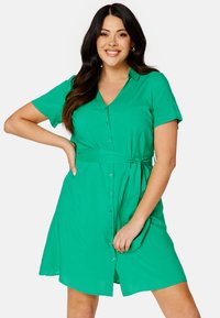 Abito camicia verde con bottoni, maniche corte, scollatura a V, cintura regolabile in vita e una silhouette A-line rilassata. Tessuto dalla trama liscia.