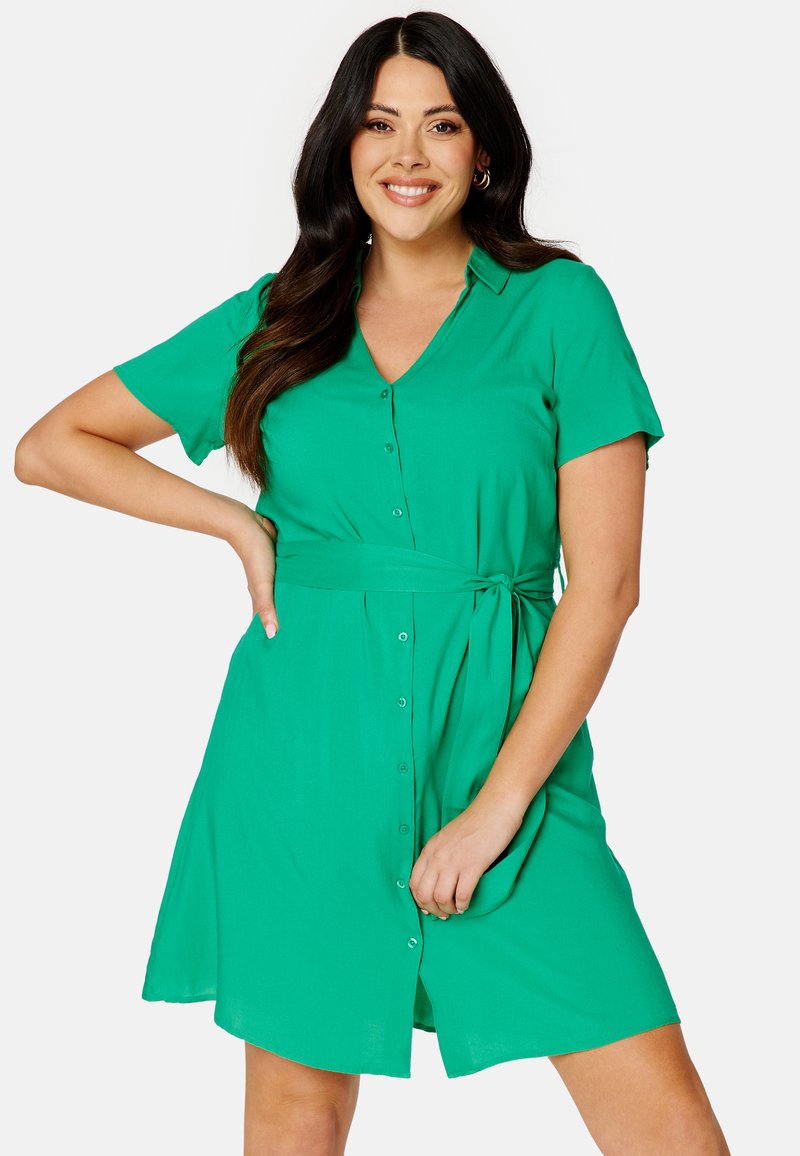 Abito camicia verde con bottoni, maniche corte, scollatura a V, cintura regolabile in vita e una silhouette A-line rilassata. Tessuto dalla trama liscia.