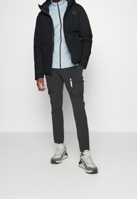 Svart isolerad jacka över en ljusblå zip-up topp, tillsammans med mörka cargo-byxor med en logotyp och grå sportskor med detaljer.