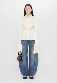 Bluză crem cu țesătură texturată și mâneci clopot supradimensionate, purtată cu blugi de denim albastri cu picioare largi și balerini negri. Posetă mică de mână de culoare închisă.