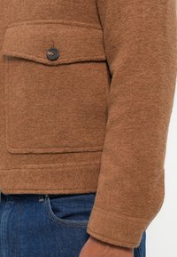 Giacca di lana marrone con tasca e dettaglio dei bottoni, caratterizzata da una texture morbida e una vestibilità su misura, abbinata a jeans in denim blu.