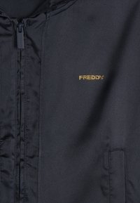 Chaqueta negra con una textura suave, que presenta una cremallera central y un pequeño logo dorado "FREDDY" en el pecho. Incluye bolsillos laterales.