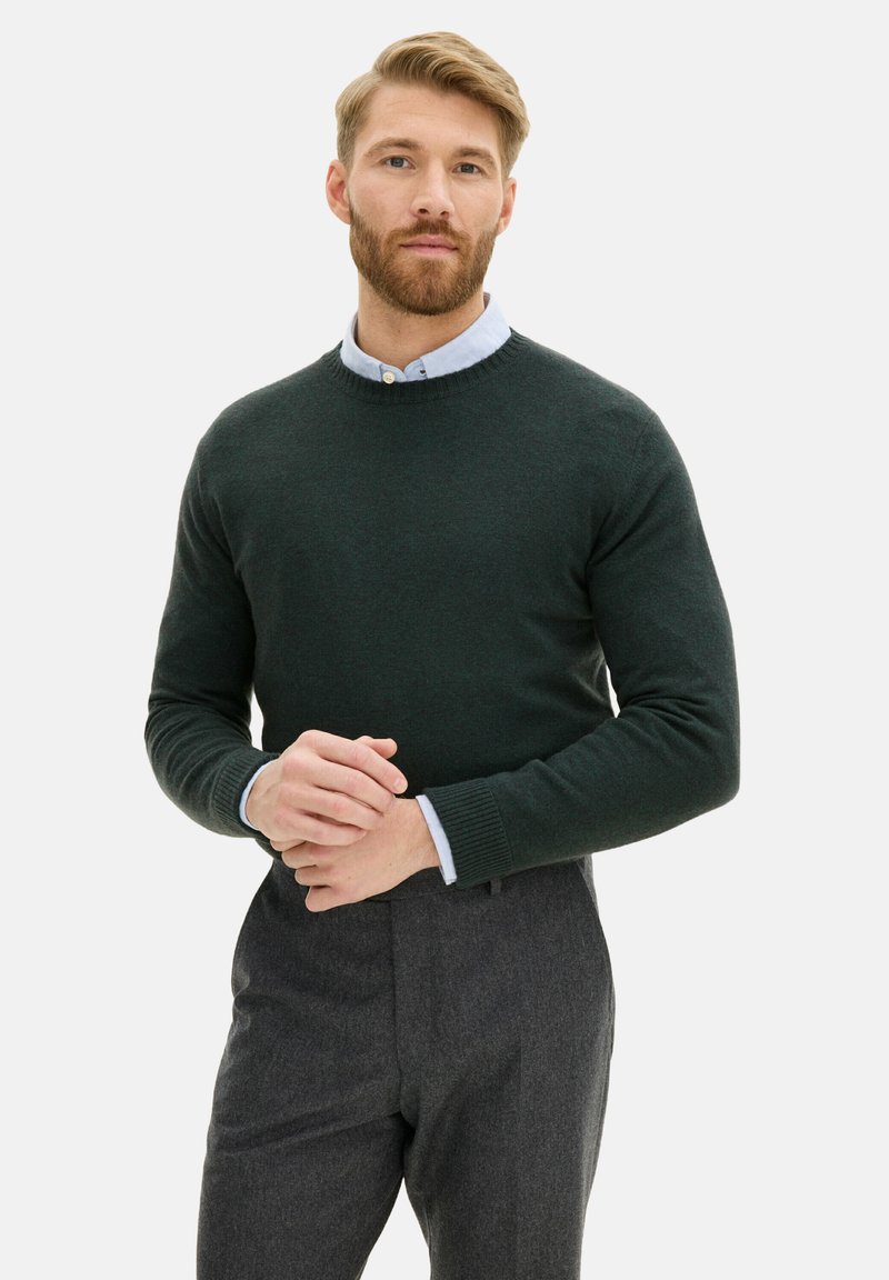 Dunkelgrüner Strickpullover mit rundem Halsausschnitt, über einem hellblauen Hemd mit Kragen getragen. Graue Anzughosen vervollständigen das Outfit.