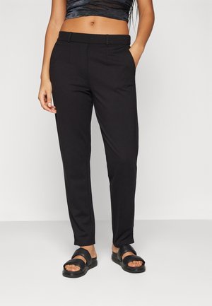 Pantalon noir fuselé avec une texture lisse, doté d'une taille plate et de poches latérales, associé à des sandales glissantes noires.