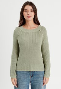 Pull en maille vert clair avec un col rond et des poignets côtelés. Présente des boutons décoratifs sur les épaules, associé à un jean bleu.