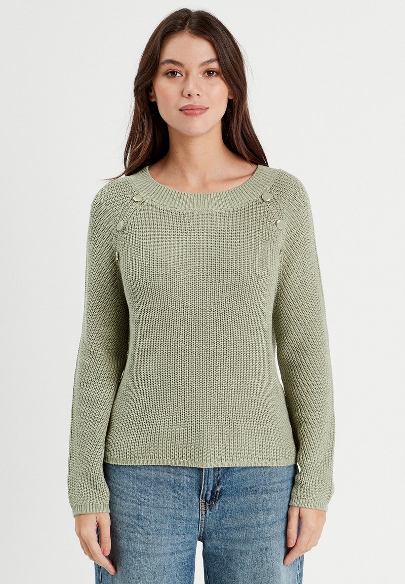 Pull en maille vert clair avec un col rond et des poignets côtelés. Présente des boutons décoratifs sur les épaules, associé à un jean bleu.