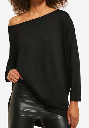 Femme portant un haut noir à épaules dénudées et manches longues, et un pantalon noir brillant en similicuir, la main tenant légèrement le haut.