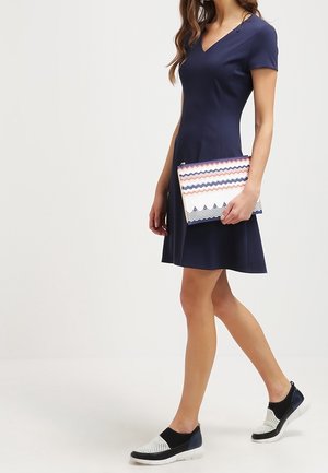 Námořnicky modré šaty s krátkým rukávem, přiléhavým pasem a rozšířenou sukní; drží vzorované psaníčko se zigzag vzory v pastelových barvách; černé slip-on boty.