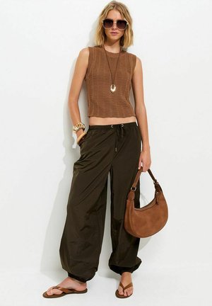 Vrouw draagt een bruin mouwloos gebreid topje, loszittende donkerbruine broek, slippers, grote zonnebril, gouden sieraden en houdt een bruine handtas vast.