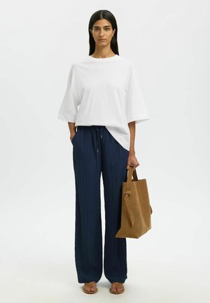 Kvinne stående iført en løs hvit skjorte, marineblå vidbukser, brune sandaler, holder en brun semsket toteveske.