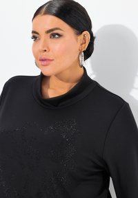 Svart sweatshirt med huva, prydd med en texturerad design av rhinestones på framsidan. Modellen bär stora silverhängen.