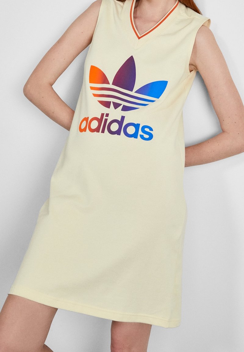 Żółta, bezrękawnikowa sukienka z dekoltem w kształcie litery V, z dużym, wielokolorowym logo Adidas i gładką bawełnianą fakturą. Wykończona pomarańczowym obszyciem.