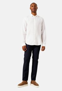 Man staat in een witte blouse met lange mouwen, donkerblauwe broek en bruine instappers tegen een effen achtergrond.