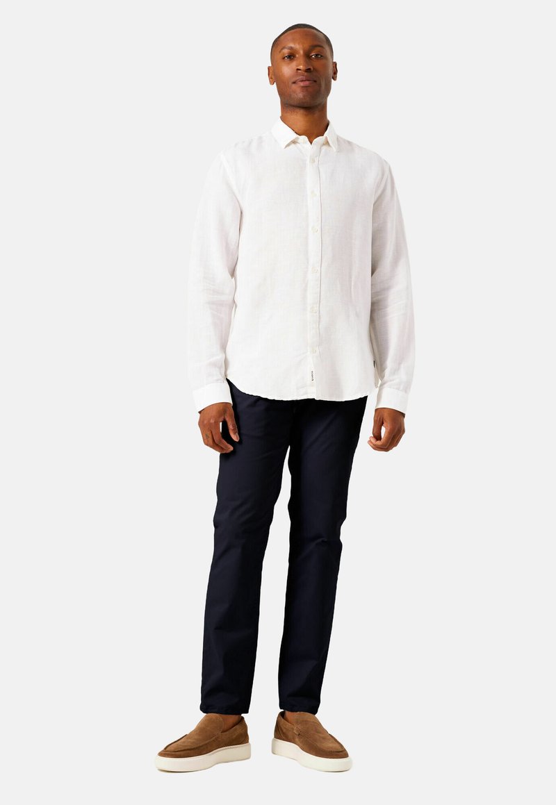 Man staat in een witte blouse met lange mouwen, donkerblauwe broek en bruine instappers tegen een effen achtergrond.