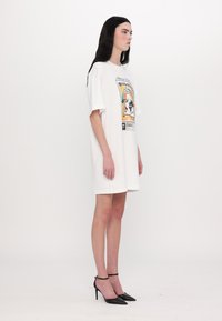 Hvit oversized t-skjortekjole med fargerik grafisk trykk. Modell iført svarte spisse hæler; rett hår og nøytral uttrykk.