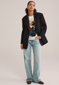 Blazer noir sur un pull blanc avec un motif de feuilles colorées, associé à un jean large bleu clair et des mocassins noirs avec des clous.