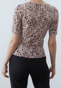 Mujer vestida con una blusa ajustada de manga corta con un patrón abstracto multicolor y pantalones negros, de pie frente a un fondo claro y liso.