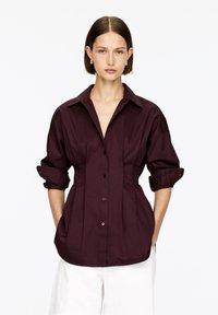 Woman with short brown hair wearing silver hoop earrings, a dark purple button-up blouse with rolled sleeves, and white pants, standing with hands in pockets.