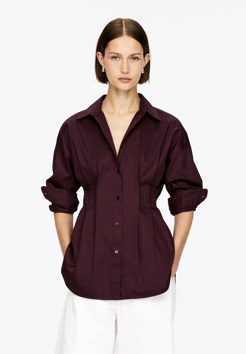 Woman with short brown hair wearing silver hoop earrings, a dark purple button-up blouse with rolled sleeves, and white pants, standing with hands in pockets.