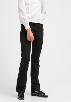 Persoon die zwarte broek met rechte pijpen, zwarte loafers met tassels en een wit blouse met lange mouwen en ruches draagt.