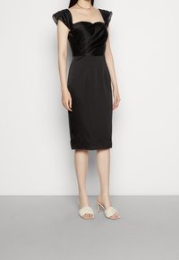 Černé saténové šaty s přiléhavým živůtkem, volnými rukávy a sukní po kolena. Spárováno s krémovými otevřenými podpatkovými mule.