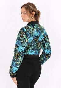 Geknipte jas van zwart stof met een blauw tropisch bladdesign en gouden kettingaccenten; ribbelkraag en -manchetten.