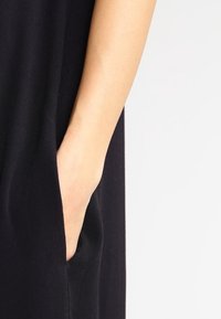 Robe noire en tissu doux, dotée d'une poche latérale et d'un design épuré et minimaliste avec une coupe décontractée. Gros plan sur une main en train d'atteindre la poche.