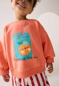Koralfarvet sweatshirt med et legende juice boks grafik med en appelsin og teksten "Squeeze the day." Stripede røde og hvide bukser fuldender looket.