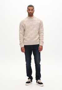 Beige kootud sviiter, millel on tekstureeritud ruuduline muster, koos tumepruunide teksade ja mustade ketsidega, millel on heledad paelad.