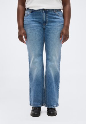 Kvinde iført lyseblå jeans med lige ben, hvid top og sorte sko, stående mod en ensfarvet baggrund.