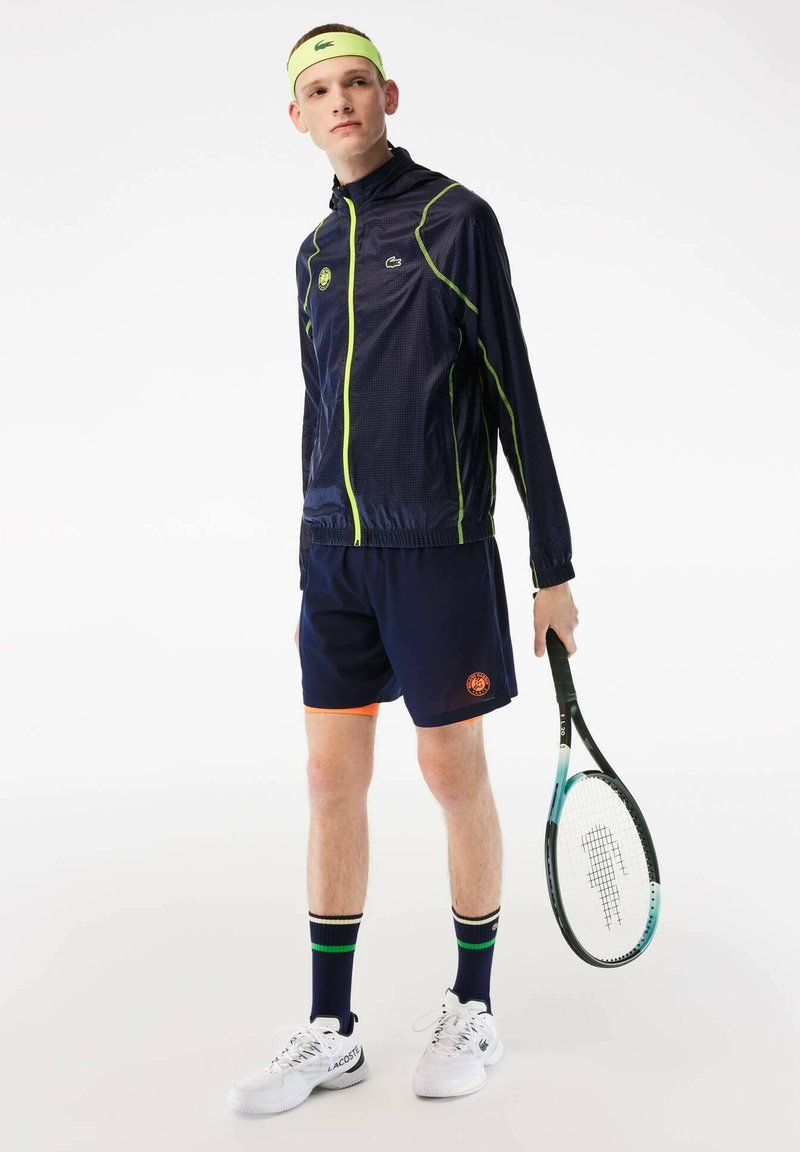 Junger männlicher Tennisspieler, der eine marineblaue Sportjacke, Shorts und ein Stirnband trägt und einen Tennisschläger in der rechten Hand hält, steht vor einem weißen Hintergrund.