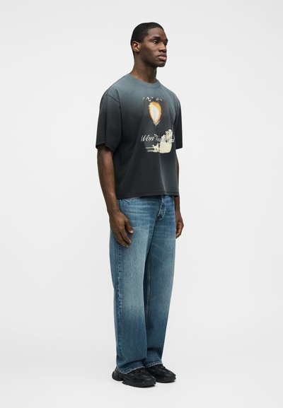 Νεαρός άνδρας που στέκεται, φορώντας μια ξεθωριασμένη μαύρη γραφική μπλούζα, μπλε τζιν με ίσια γραμμή και μαύρα αθλητικά παπούτσια σε απλό λευκό φόντο.